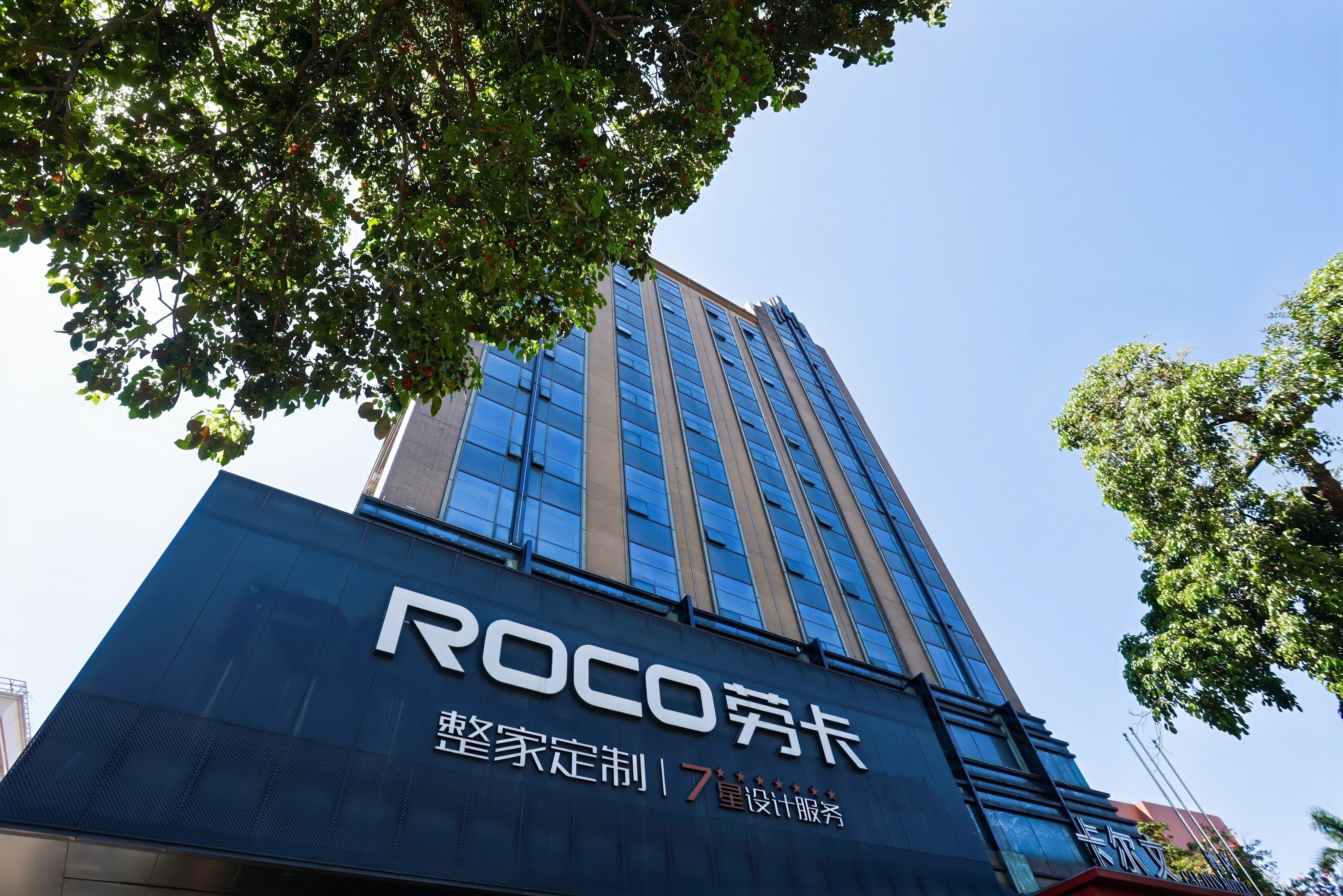 户外仰视视角的劳卡（ROCO）品牌大楼外观，主体为现代商业建筑，配备蓝色玻璃幕墙与深色金属质感招牌。招牌醒目展示“ROCO 劳卡”品牌标识及“整家定制 | 7星